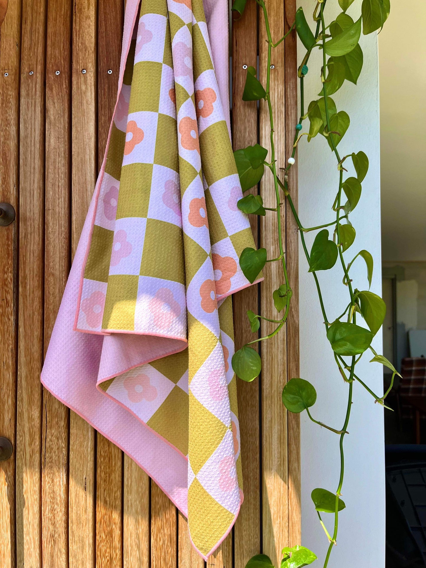 A pink sand free towel with a geometric pattern featuring palm leaves, draped over a wooden surface with a plant in the background.