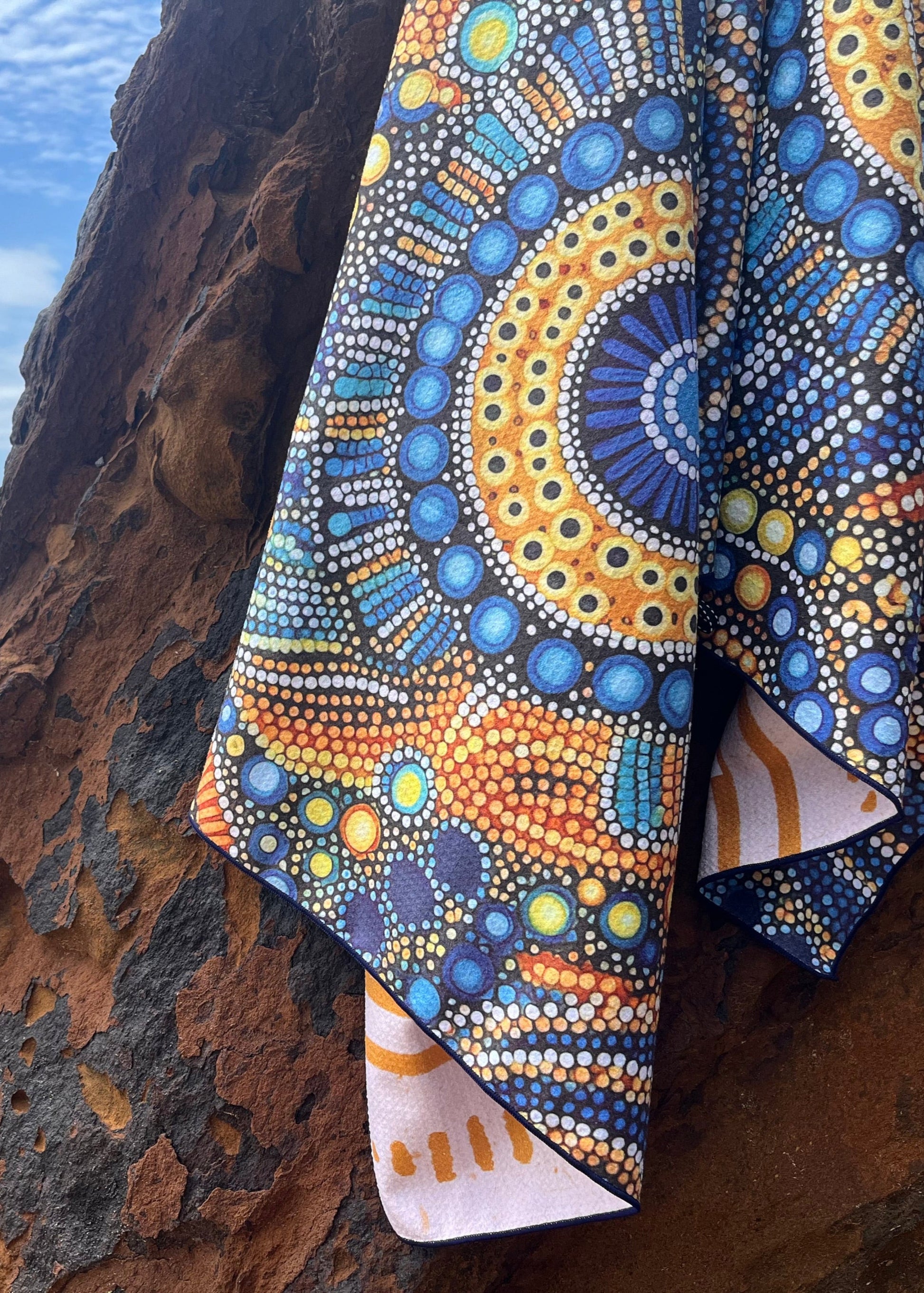 A multicolored sand freetowel with a geometric pattern is draped over a rocky outcrop with a beach and ocean in the background.