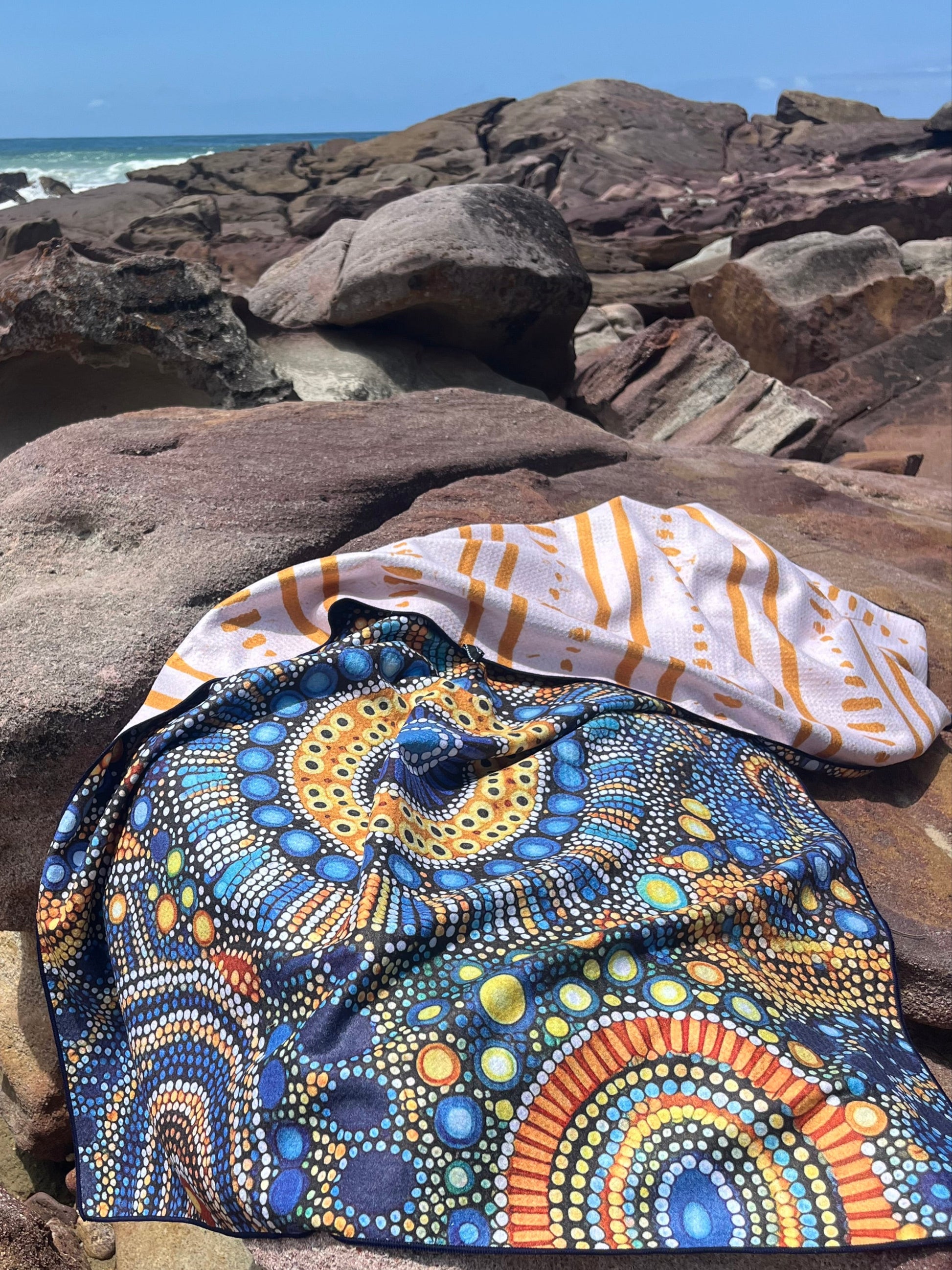 A multicolored towel with a geometric pattern is laid out on a rocky surface with a beach and rocks in the background.