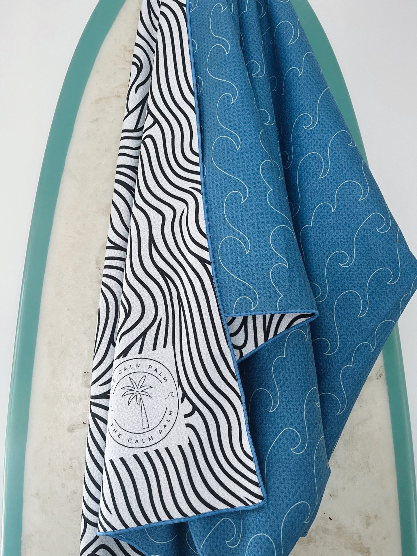 A beach towel with a wave pattern design, featuring a built-in headrest pocket, draped over a surfboard.