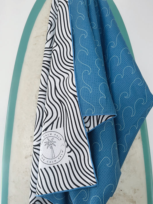A beach towel with a wave pattern design, featuring a built-in headrest pocket, draped over a surfboard.