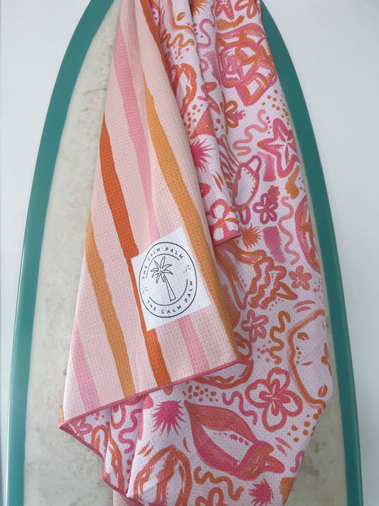 A beach towel with a mix of striped and floral patterns, predominantly in pink and orange shades, featuring a built-in headrest pocket and a brand logo on a white patch.