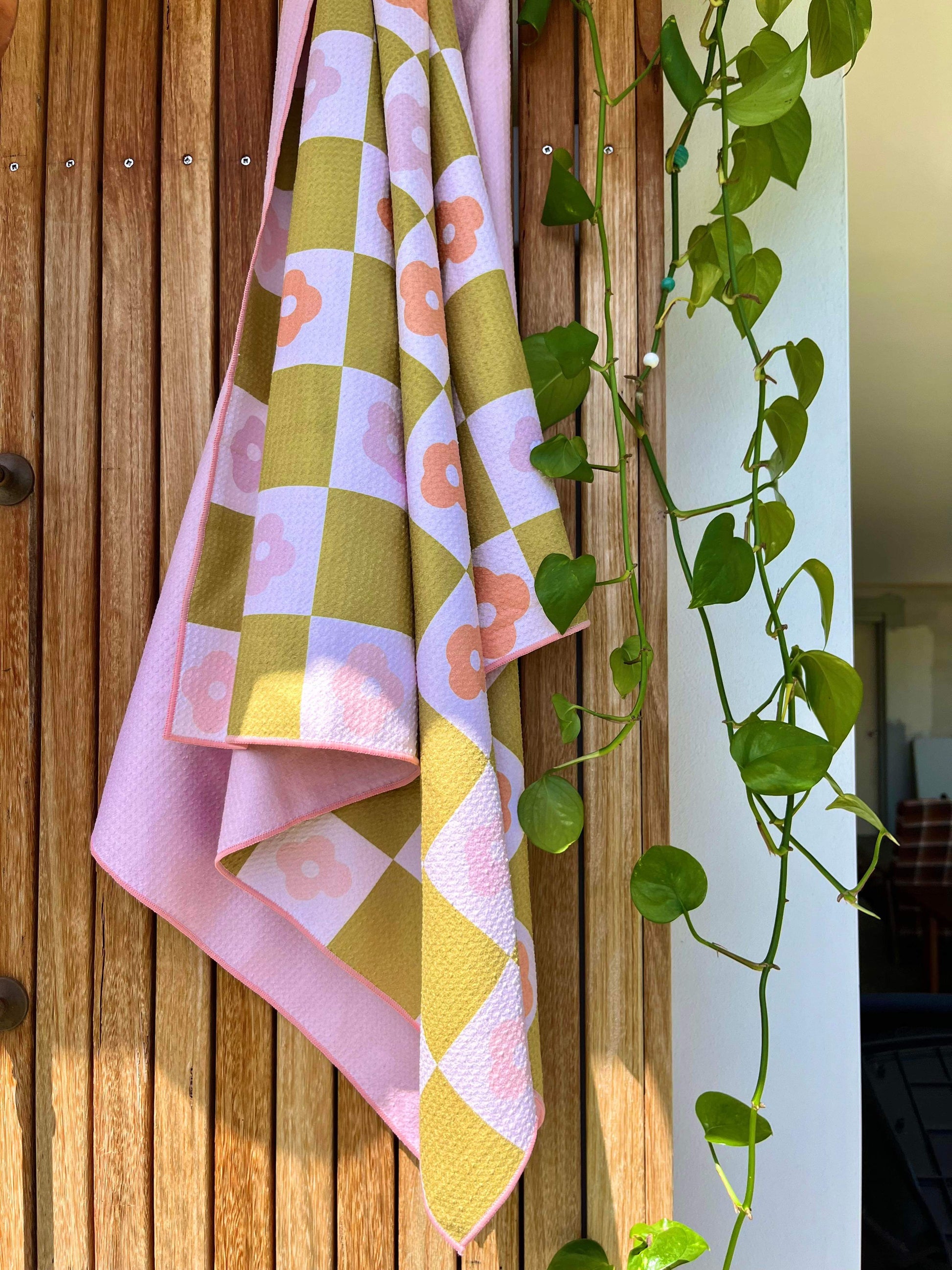 A pink sand free towel with a geometric pattern featuring palm leaves, draped over a wooden surface with a plant in the background.