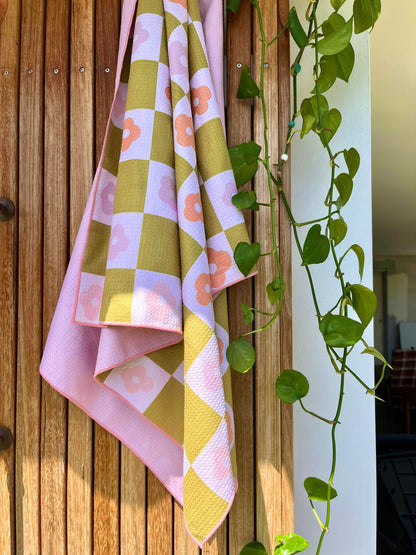 A pink sand free towel with a geometric pattern featuring palm leaves, draped over a wooden surface with a plant in the background.