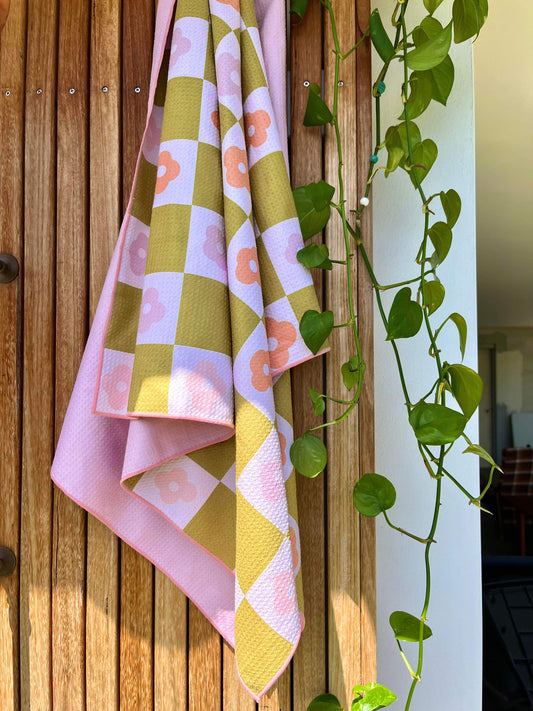 A pink sand free towel with a geometric pattern featuring palm leaves, draped over a wooden surface with a plant in the background.