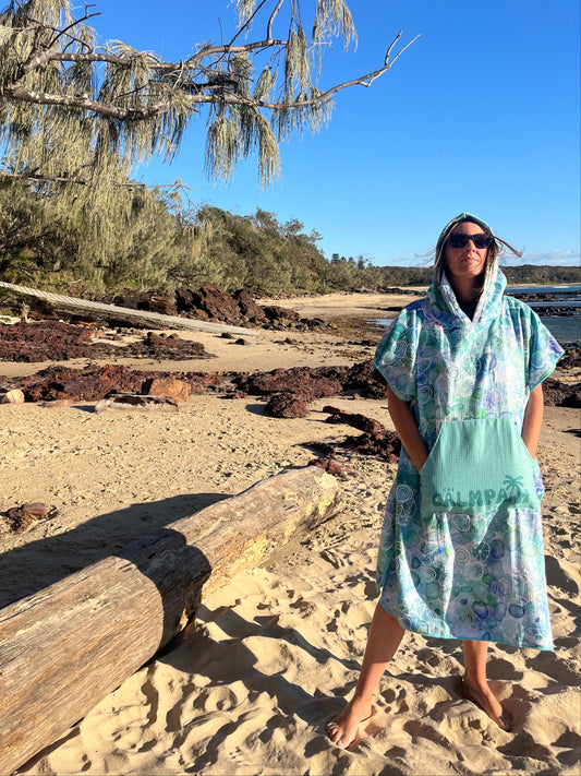 Person in a light blue hooded beach towel standing on a rocky cliff with trees and clear sky in the background